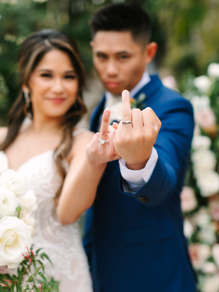 Bride and groom smiling, showing wedding rings with a playful gesture.