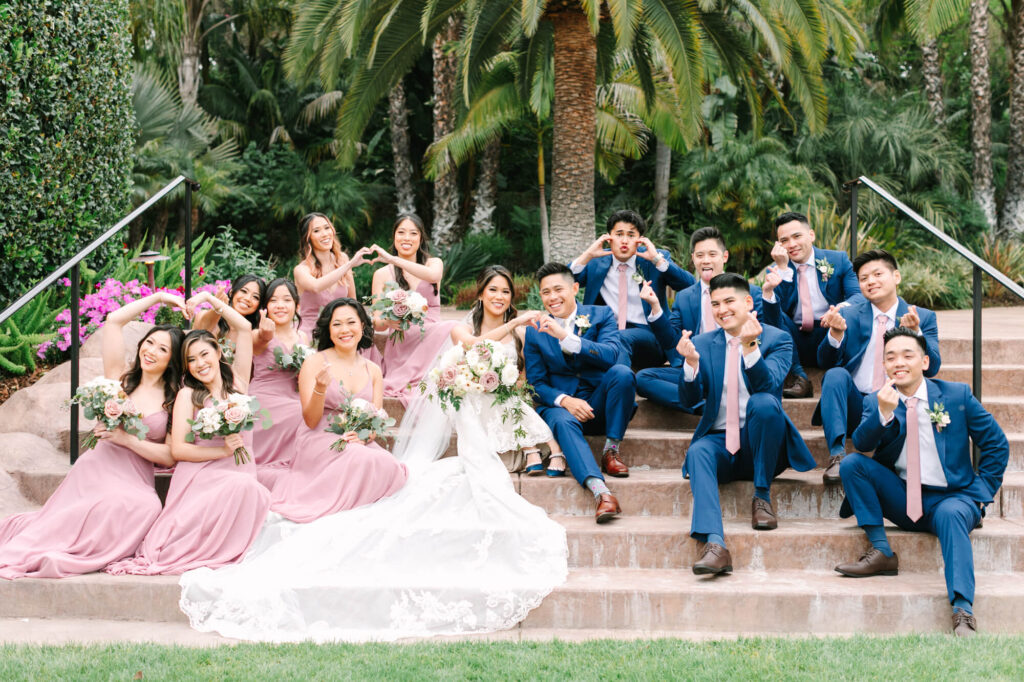 Bridal party on steps, bridesmaids in pink dresses, groomsmen in blue suits with pink ties, all making heart shapes with hands, lush greenery backdrop.