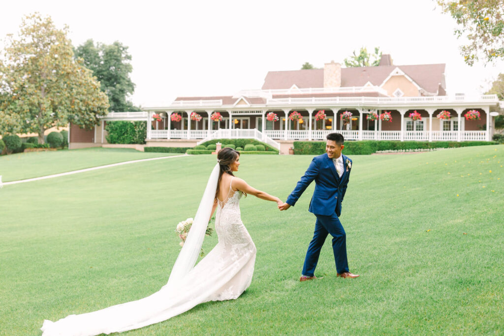 A bride in a flowing white gown and veil holds hands with a groom in a blue suit on a lush green lawn, leading to a grand house adorned with flowers.