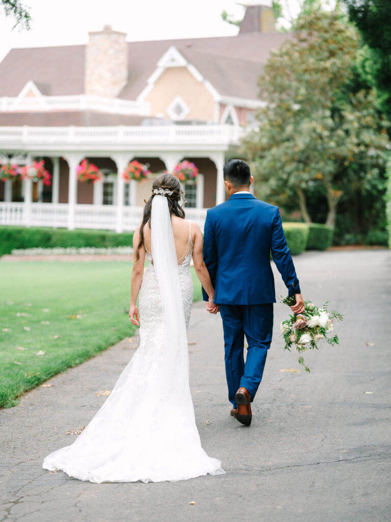 Bride and groom hold hands, walking away on a path. The bride wears a white gown, and the groom wears a blue suit, with a bouquet.