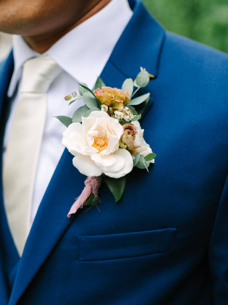 A close-up of a man in a blue suit with a cream-colored tie. A boutonniere with white roses and greenery adorns his jacket.