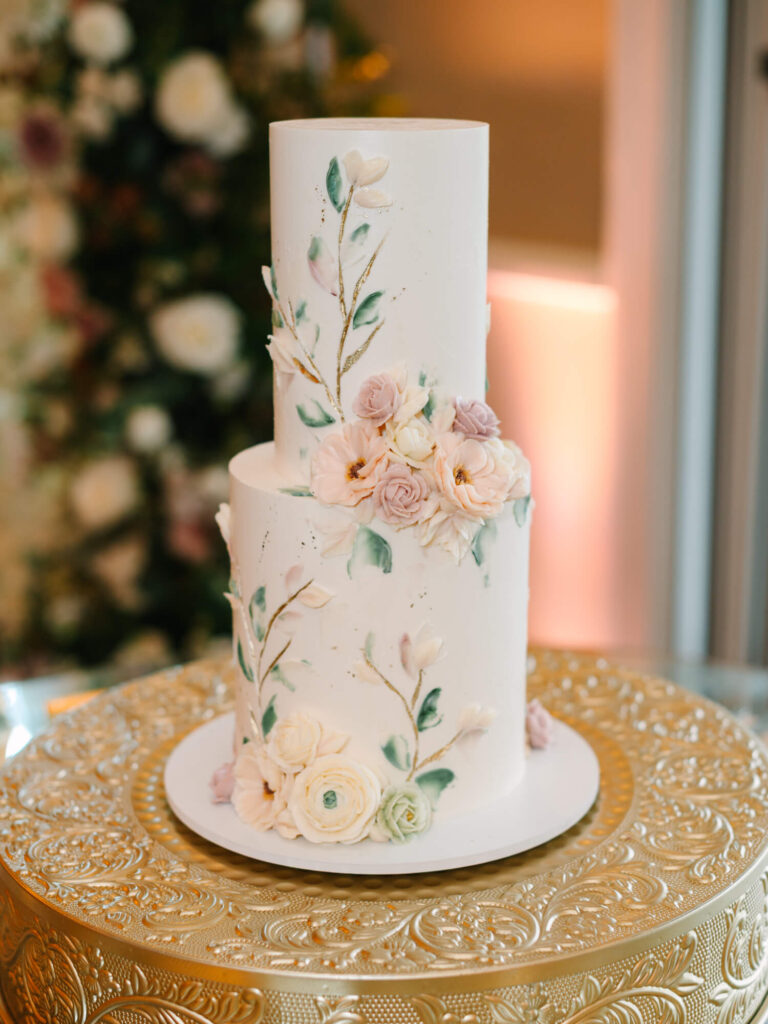 Two-tier white cake with pink and white floral decorations, placed on an ornate gold platter.