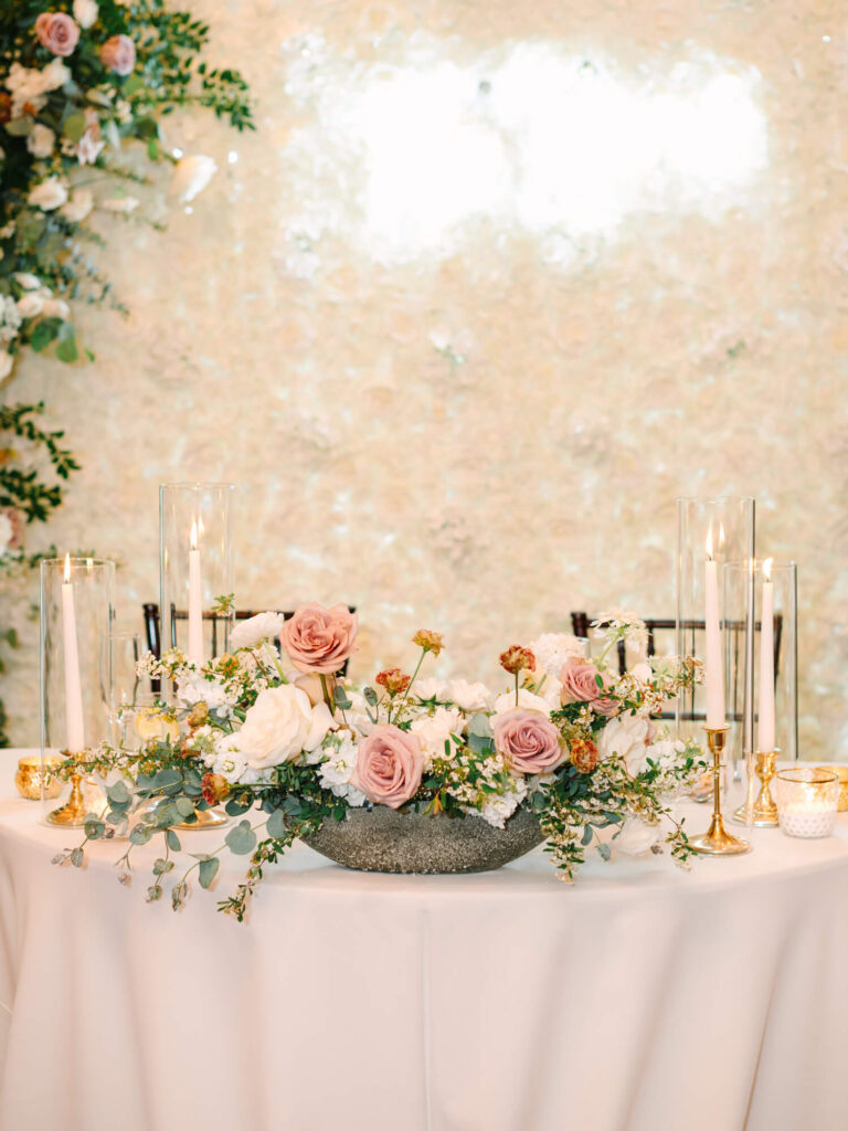 Elegant table setting with a floral centerpiece featuring pink and white roses, greenery, and candles in glass holders.