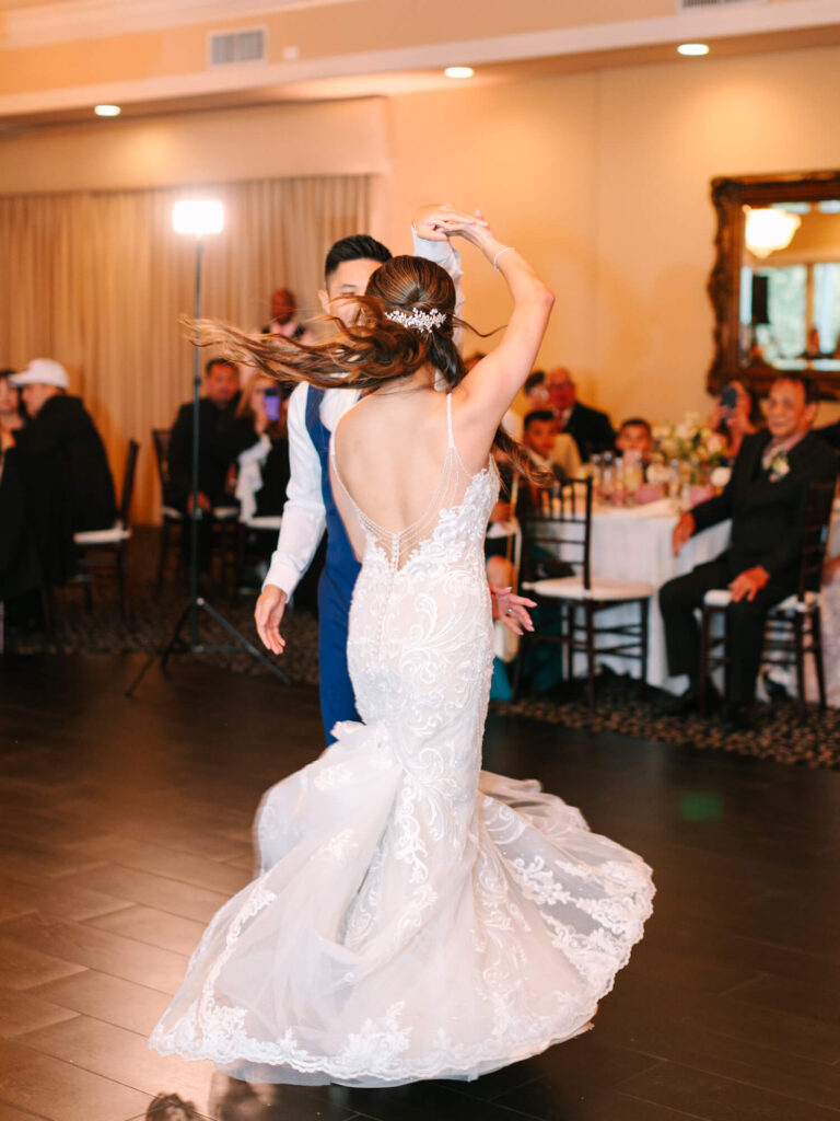 A bride and groom dance joyfully, her lace dress swirling gracefully. Guests seated at round tables watch with smiles under warm lighting.