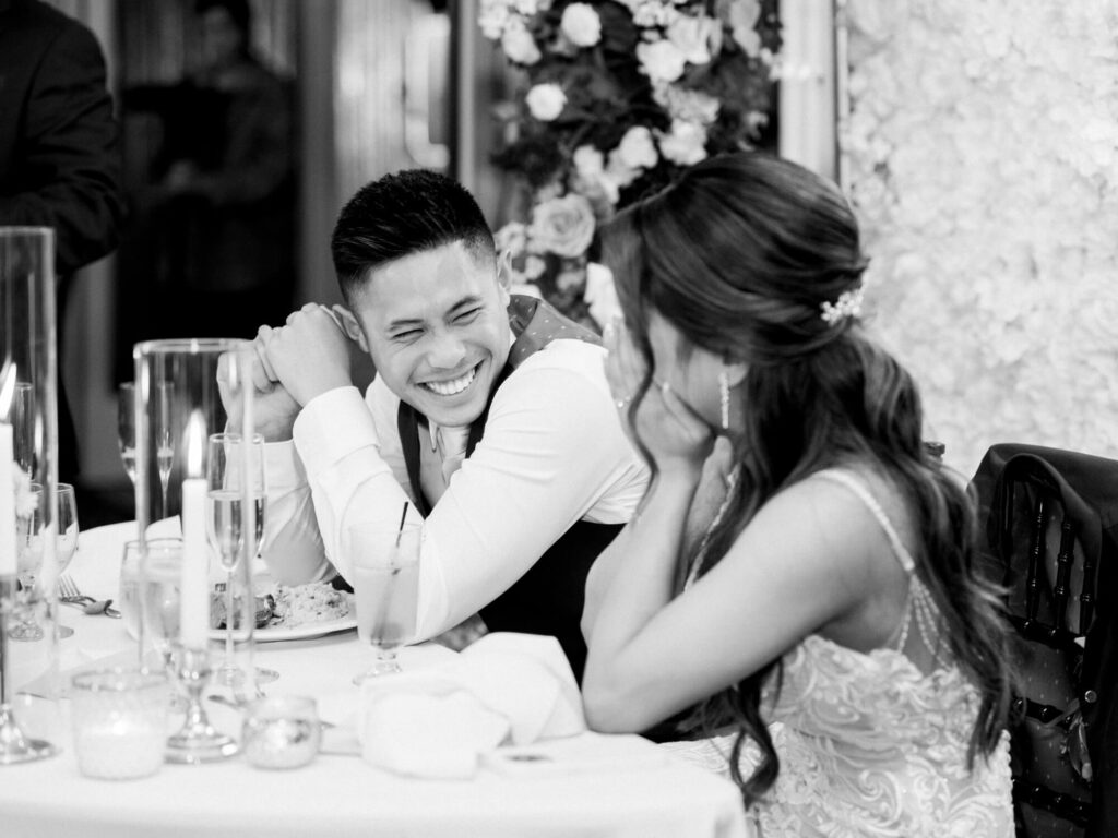 A couple sits at a wedding reception table, laughing joyfully.