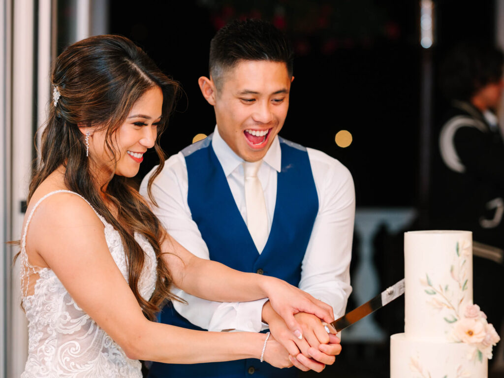 A joyful couple cuts a white wedding cake adorned with floral designs.