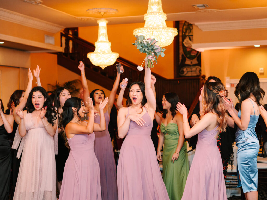 Bridesmaids in lilac dresses cheer as one catches the bouquet at a wedding reception.