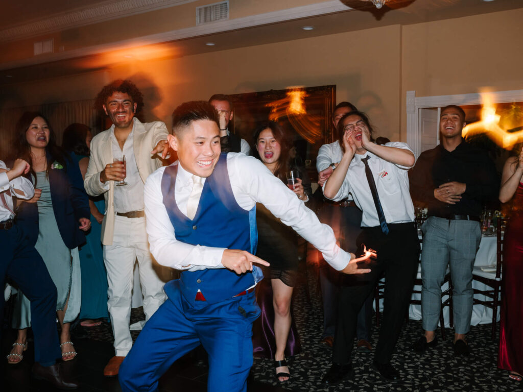 Groom in a blue suit energetically dances at a party, surrounded by cheering friends.
