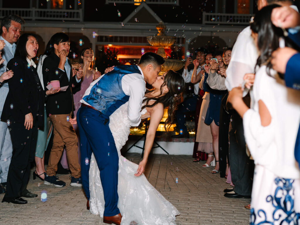 A couple shares a joyful kiss during their wedding, surrounded by guests blowing bubbles.
