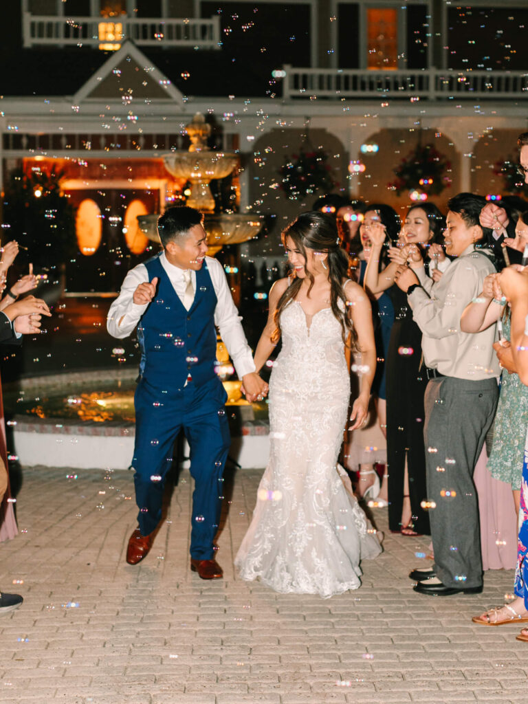 Bride and groom in formal attire walk hand in hand through a crowd blowing bubbles.