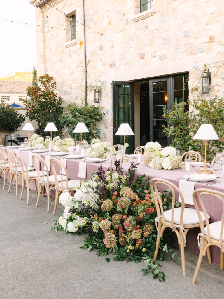 Elegant outdoor table setup with white chairs, lilac tablecloths, and white flowers.