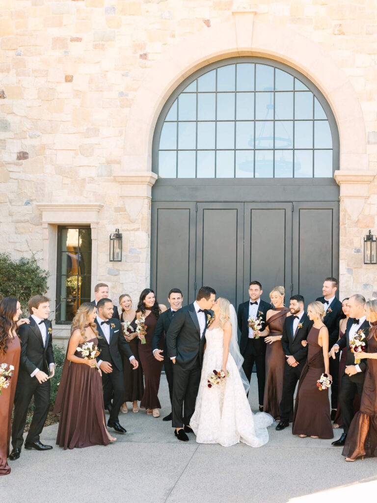 A joyful wedding party gathers around a kissing bride and groom in front of a grand stone building. Bridesmaids wear dark brown dresses, holding bouquets.