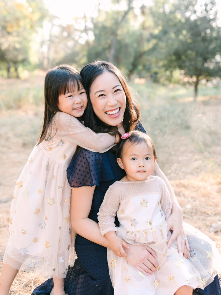 A joyful woman is outdoors with two young children. She is smiling broadly, holding one child, while the other hugs her.