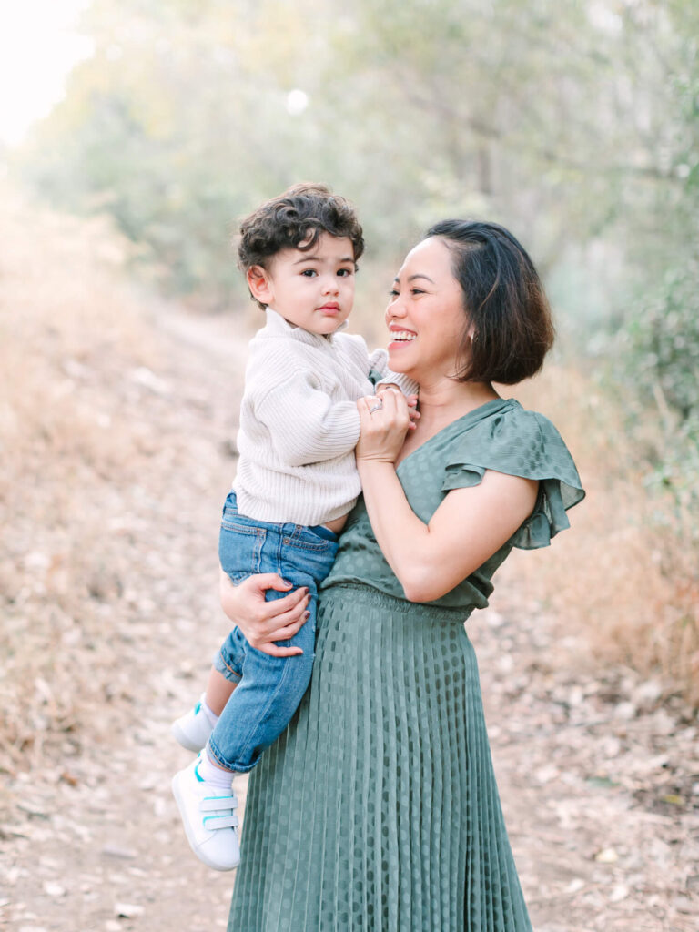 A woman in a green dress joyfully holds a young child wearing a sweater and jeans on a leafy path.