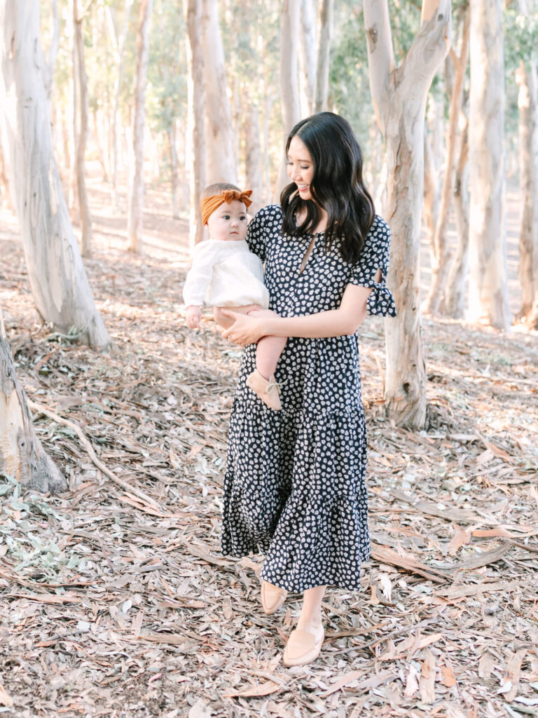 A woman in a polka-dot dress holds a baby wearing a white outfit and orange headband in a sunlit forest