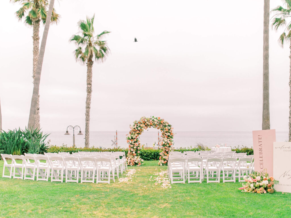 Oceanfront wedding setup with white chairs facing a floral arch, flanked by palm trees. Overcast sky and calm sea.