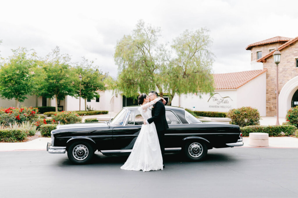 A couple in wedding attire embraces in front of a classic black car.