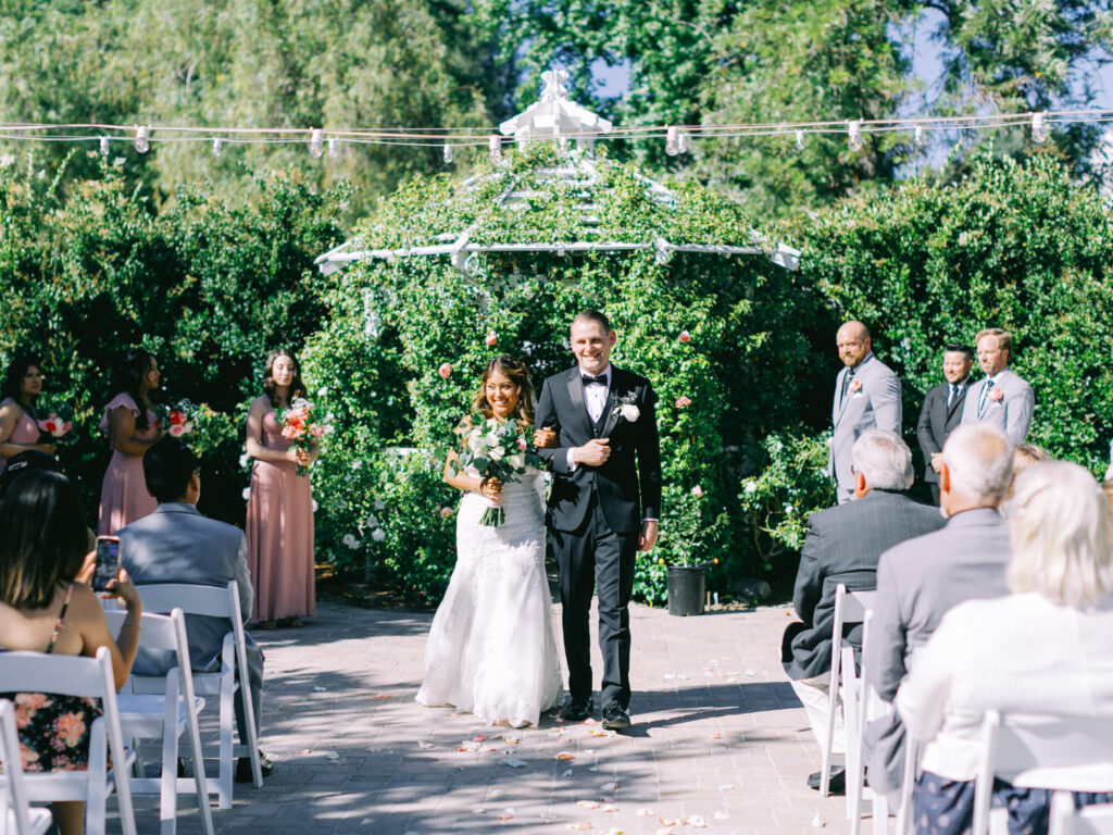 A bride in a white dress and a groom in a black tuxedo walk down an outdoor aisle, surrounded by greenery and guests.