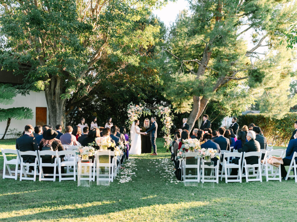 Outdoor wedding ceremony in a garden. A couple stands under a floral arch, surrounded by trees and guests seated on white chairs.