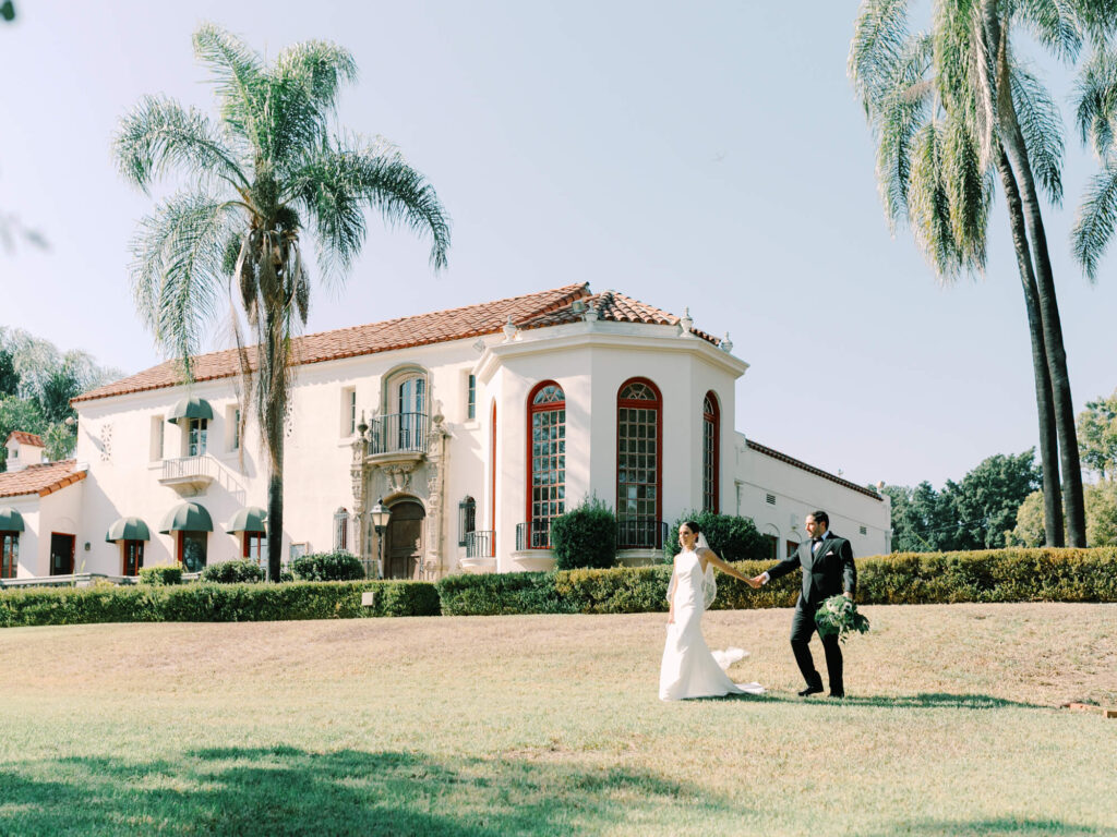 A bride and groom walk hand in hand on a lush lawn in front of a grand, white villa with red-tiled roof and large windows, surrounded by tall palm trees.