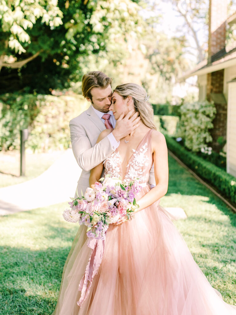 A couple embraces in a garden, the woman in a blush pink gown holding a bouquet of flowers.