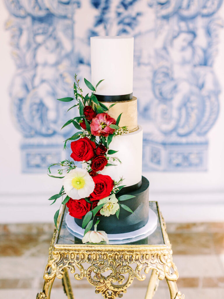 Elegant four-tier cake with white, gold, and black layers, adorned with red and white flowers.