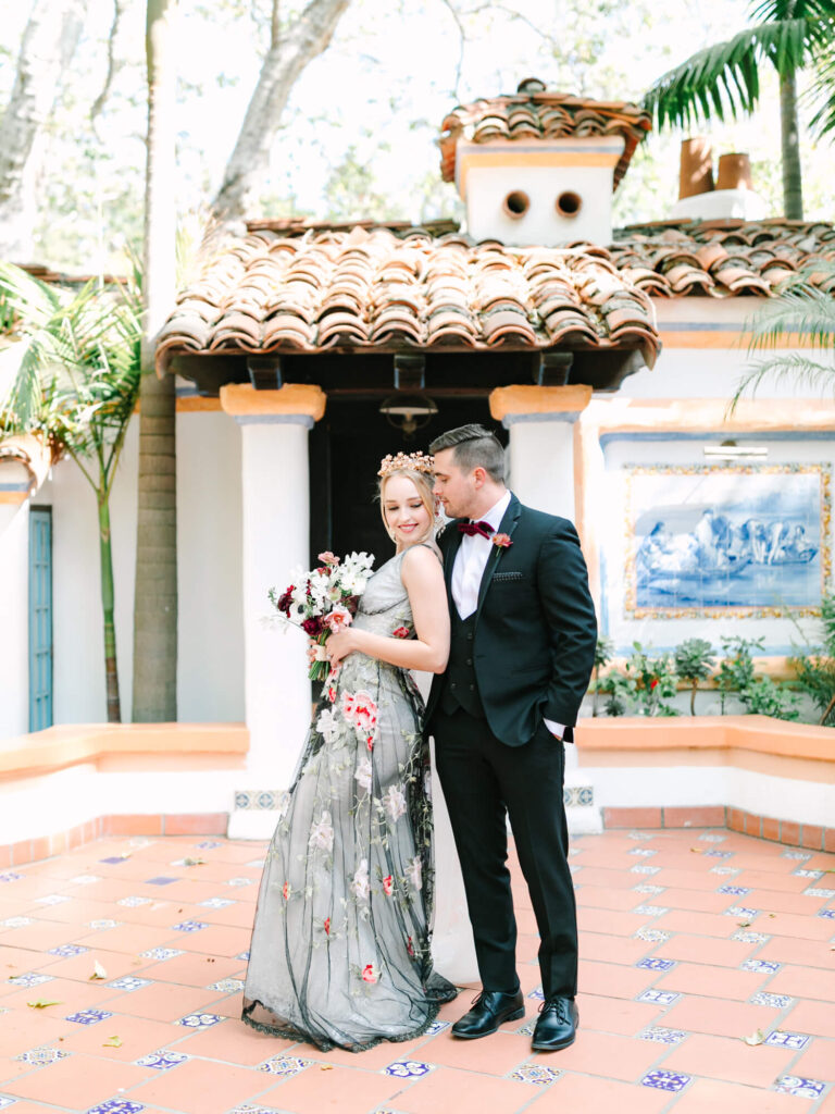 A couple stands in a courtyard with Spanish-style architecture. The woman holds a floral bouquet and wears a light dress with a floral pattern.
