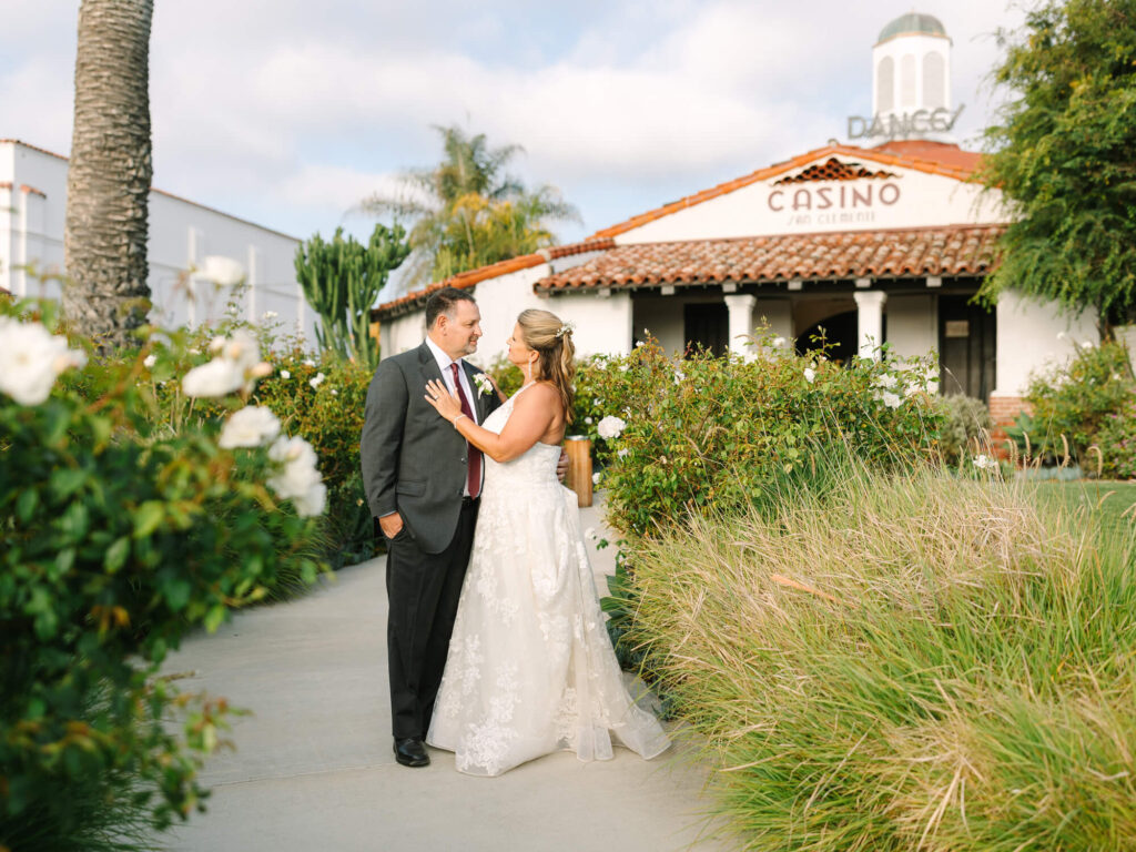 A couple in wedding attire shares an intimate moment on a garden path. They're in front of a historic building with a "Casino" sign, surrounded by lush greenery.