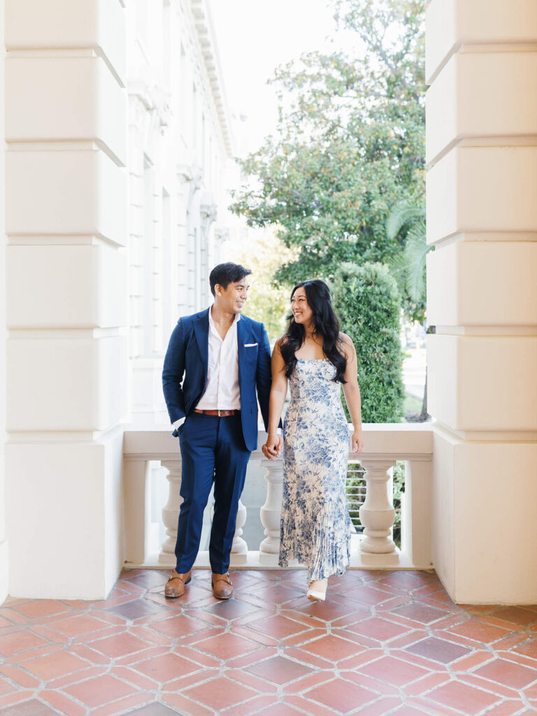 A couple walks hand in hand, smiling at each other. The man wears a blue suit, and the woman a floral dress.