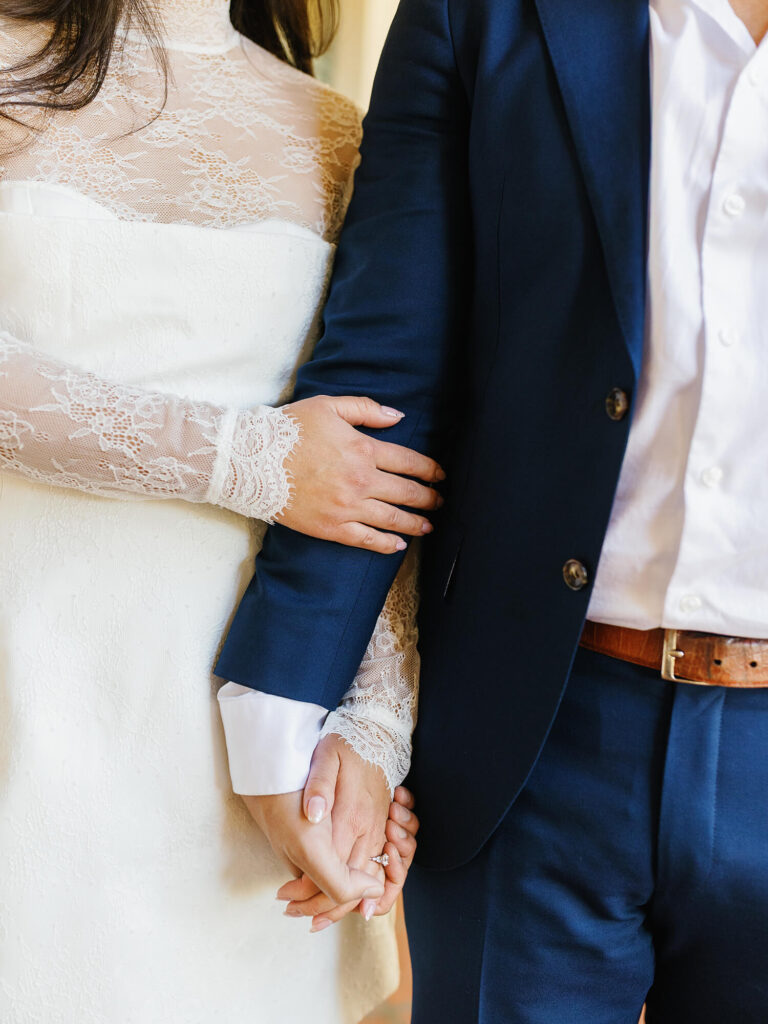 A bride in a lace dress lovingly holds a man's arm, wearing a navy suit, while their hands are clasped.