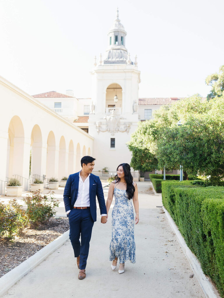 A couple walks hand smiling at each other. They are in a garden with trimmed hedges and a tall, elegant building behind them.