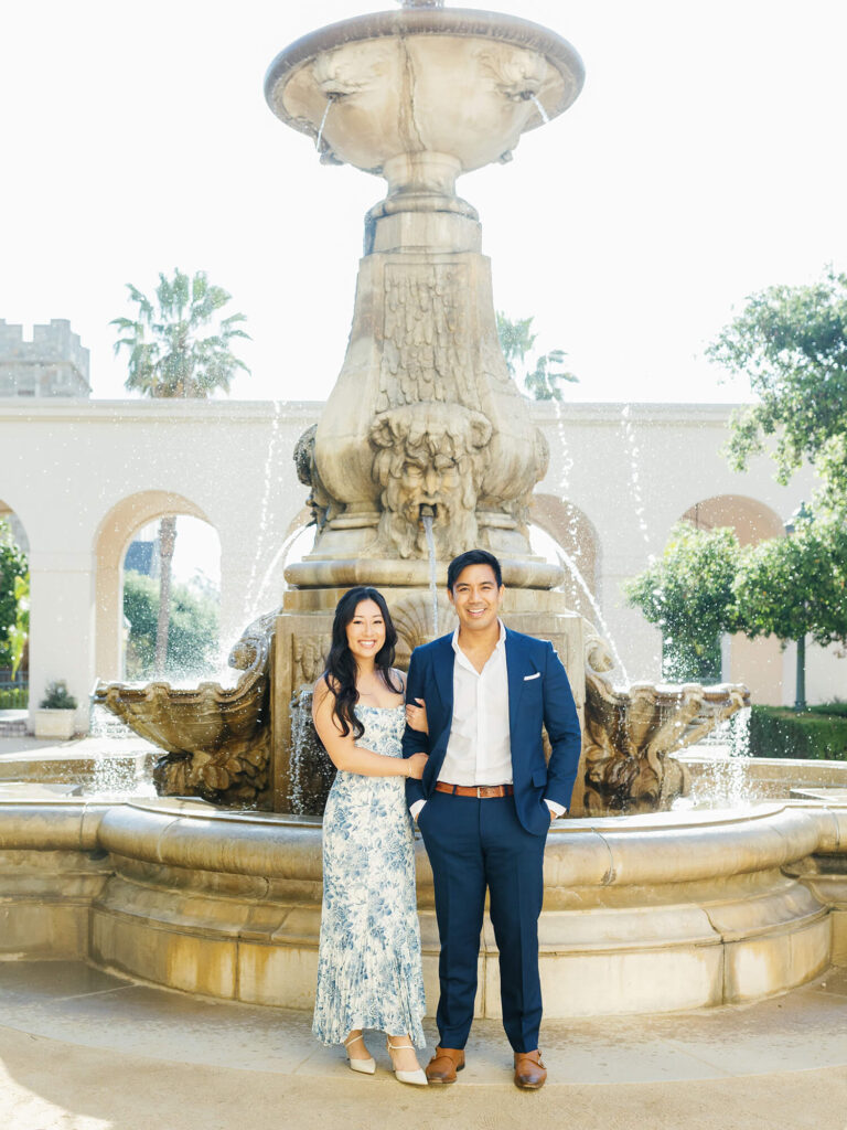 A couple stands smiling in front of an ornate stone fountain, surrounded by arches and greenery.