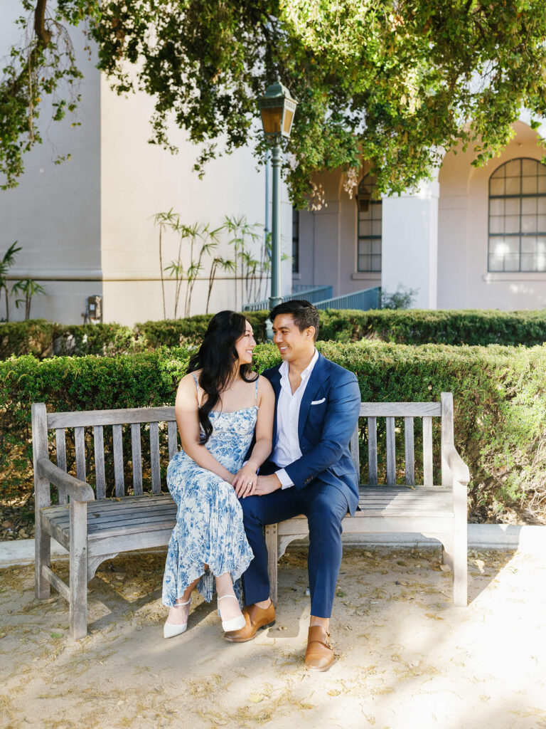 A couple sits closely on a wooden bench under a tree. The woman wears a blue floral dress, and the man is in a blue suit. They smile at each other warmly.