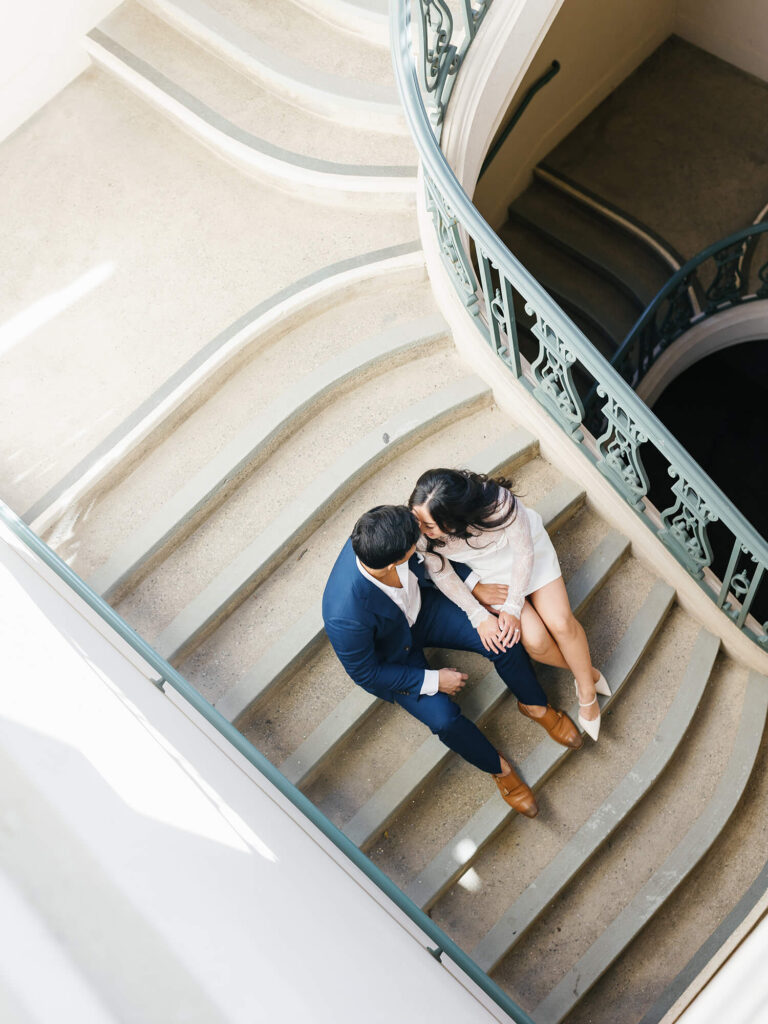 A couple sits closely on a curved staircase, sharing a quiet moment. The man wears a blue suit, the woman a white dress.