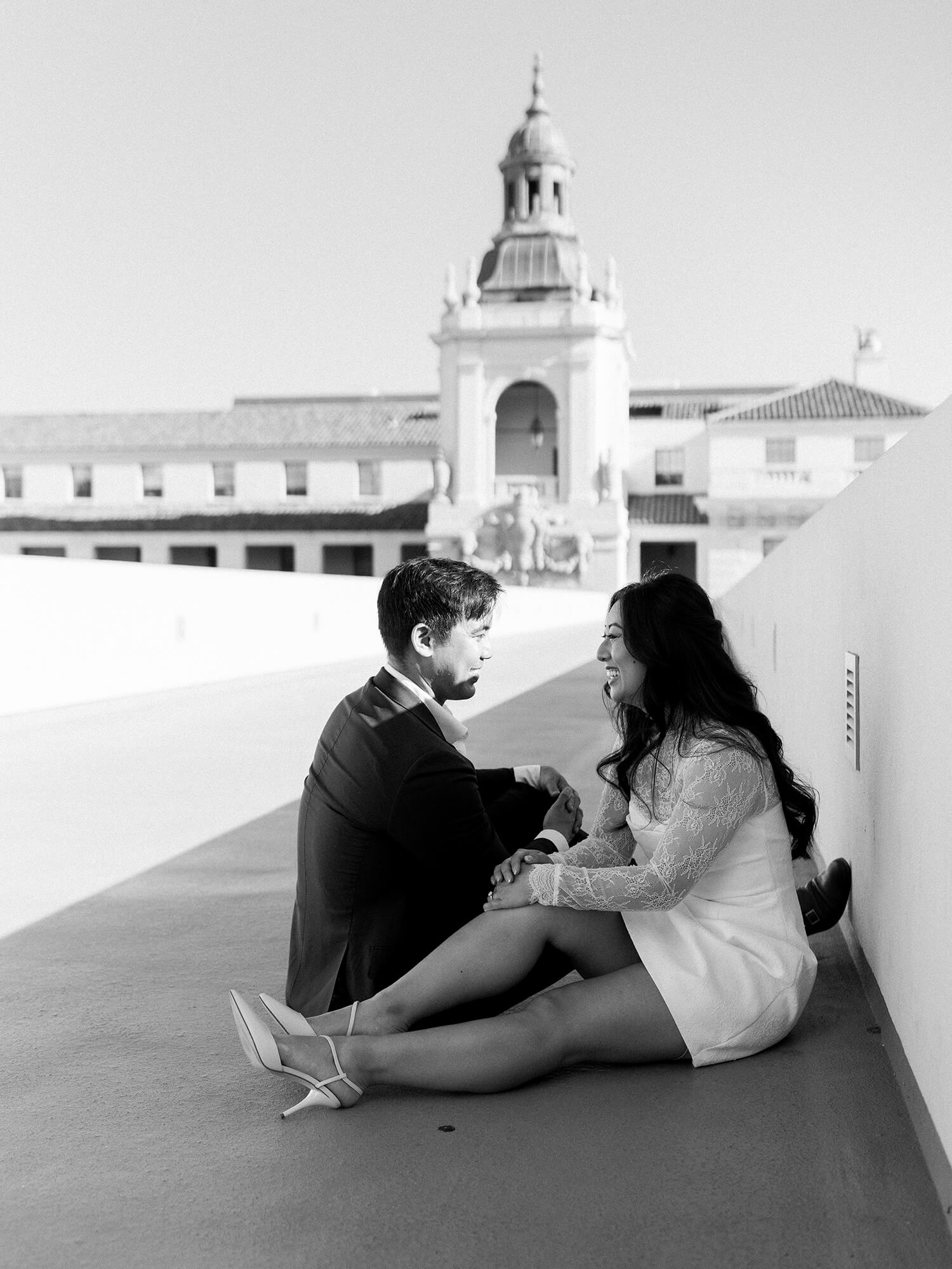 A couple sits on a rooftop with a historic building in the background. They are smiling at each other.