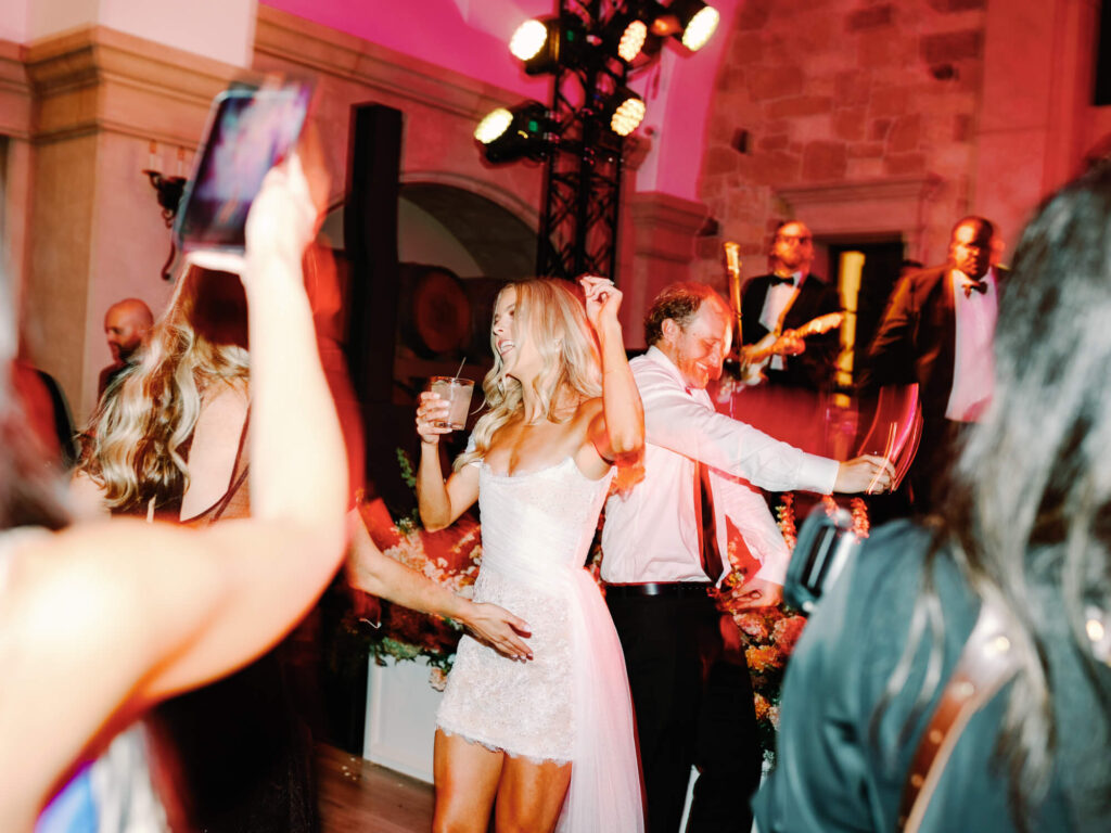 A joyful bride in a white dress dances with guests at a lively indoor wedding reception. People are smiling, and a band plays in the background.