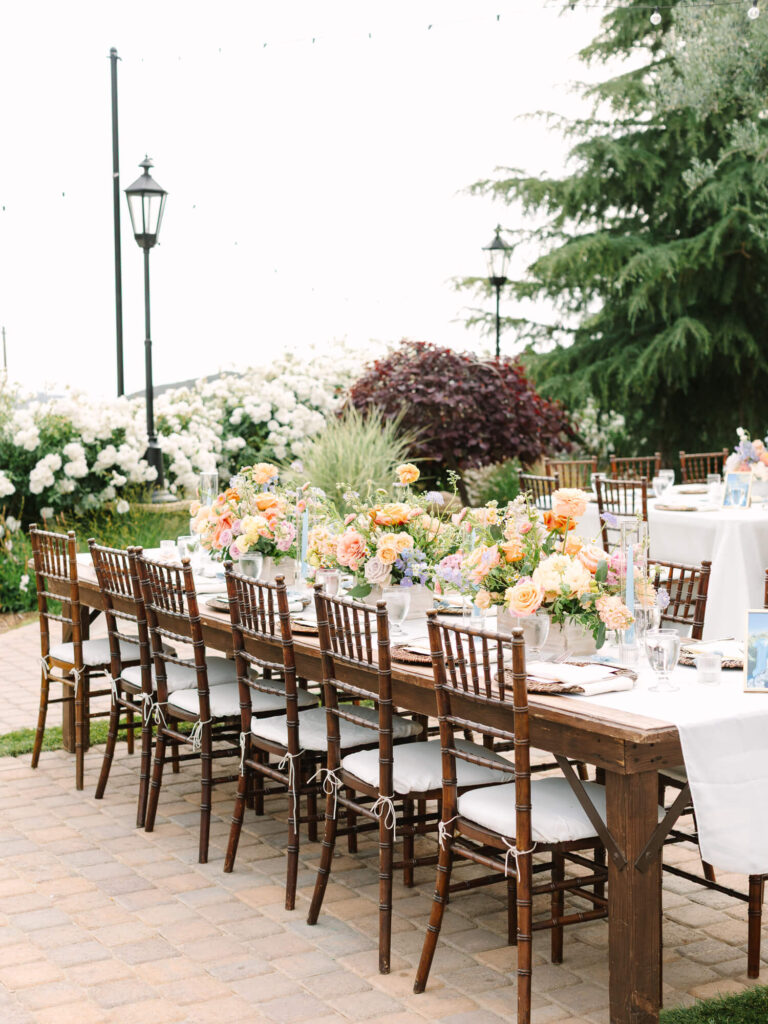 Elegant outdoor dining setup with a long wooden table, adorned with vibrant flower arrangements and surrounded by brown chairs.