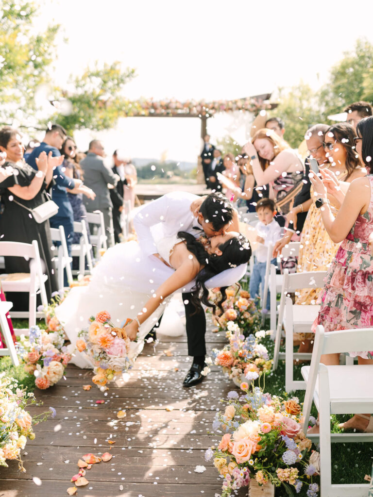 A newlywed couple shares a joyful, romantic dip kiss down an outdoor aisle, surrounded by applauding guests and vibrant floral arrangements.