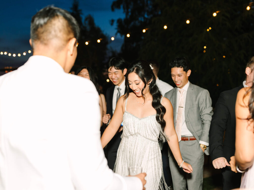 A joyful group dances at an outdoor evening event. Bride in a silver fringe dress smiles, surrounded by people in formal wear and string lights.