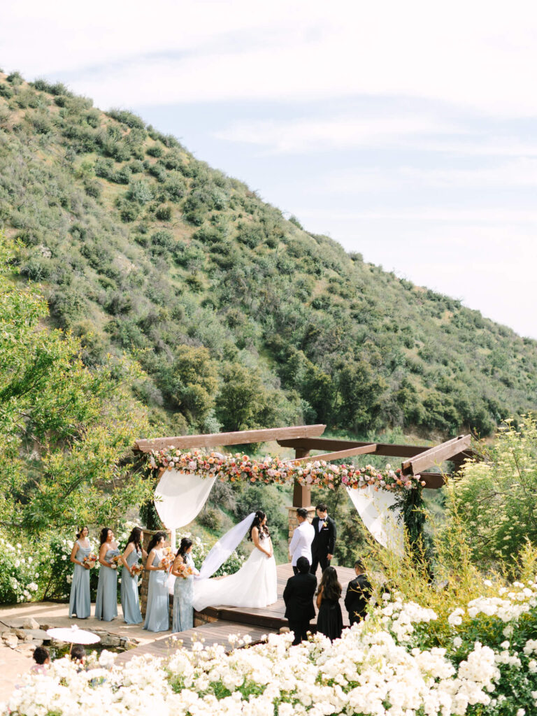 Bride and groom exchange vows under a floral arch on a wooden deck, surrounded by bridal party. Lush green hills and white roses frame the scene.