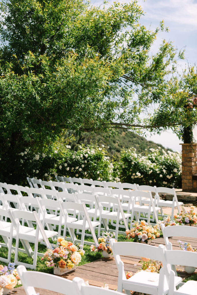 Rows of white chairs are set up outdoors for a wedding ceremony, surrounded by lush greenery and colorful flower arrangements, under a sunny sky.