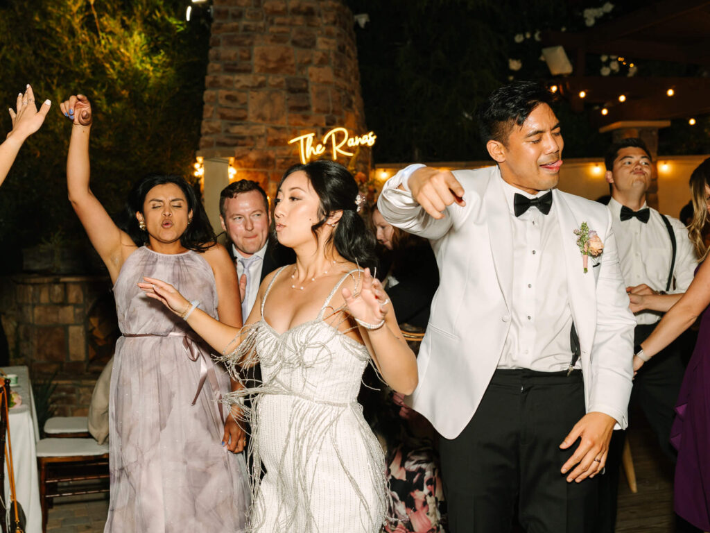 A lively wedding reception with guests joyfully dancing. People are dressed in formal attire. Warm lighting and a sign reading "The Ranas" in the background.