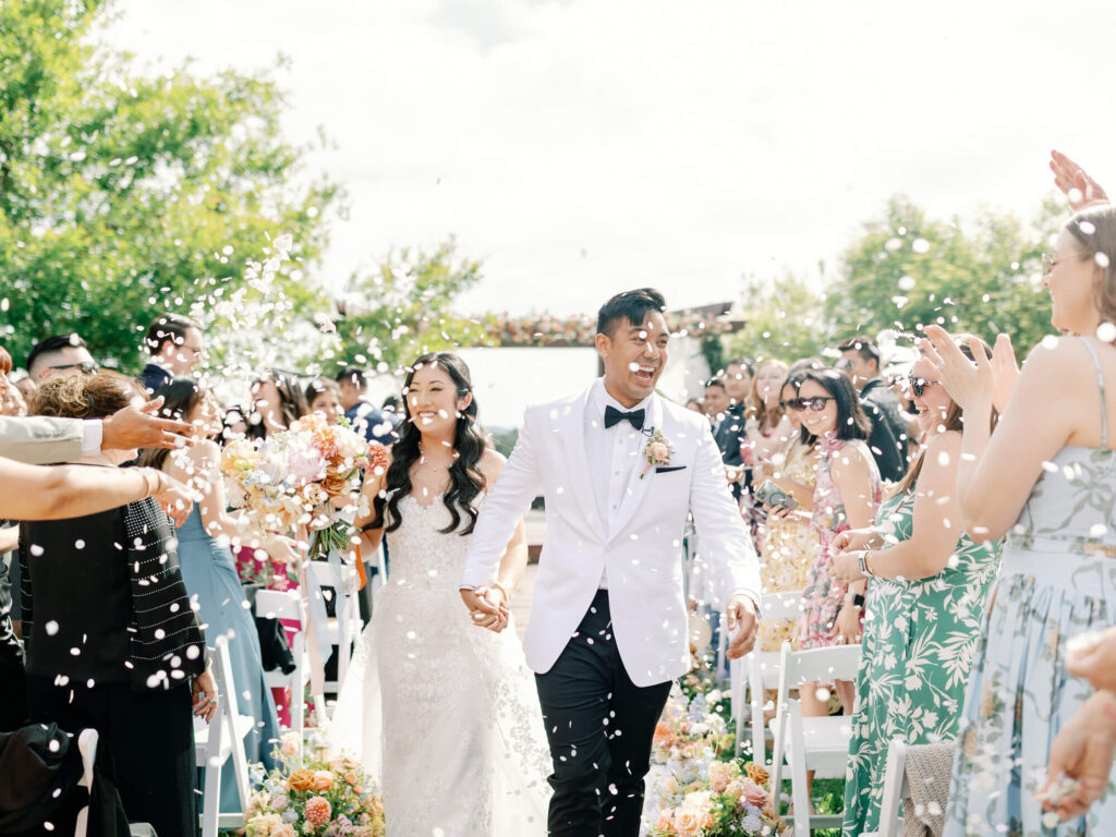 A joyful couple walks down the aisle bathed in sunlight, amidst a shower of petals. Guests clap and smile.