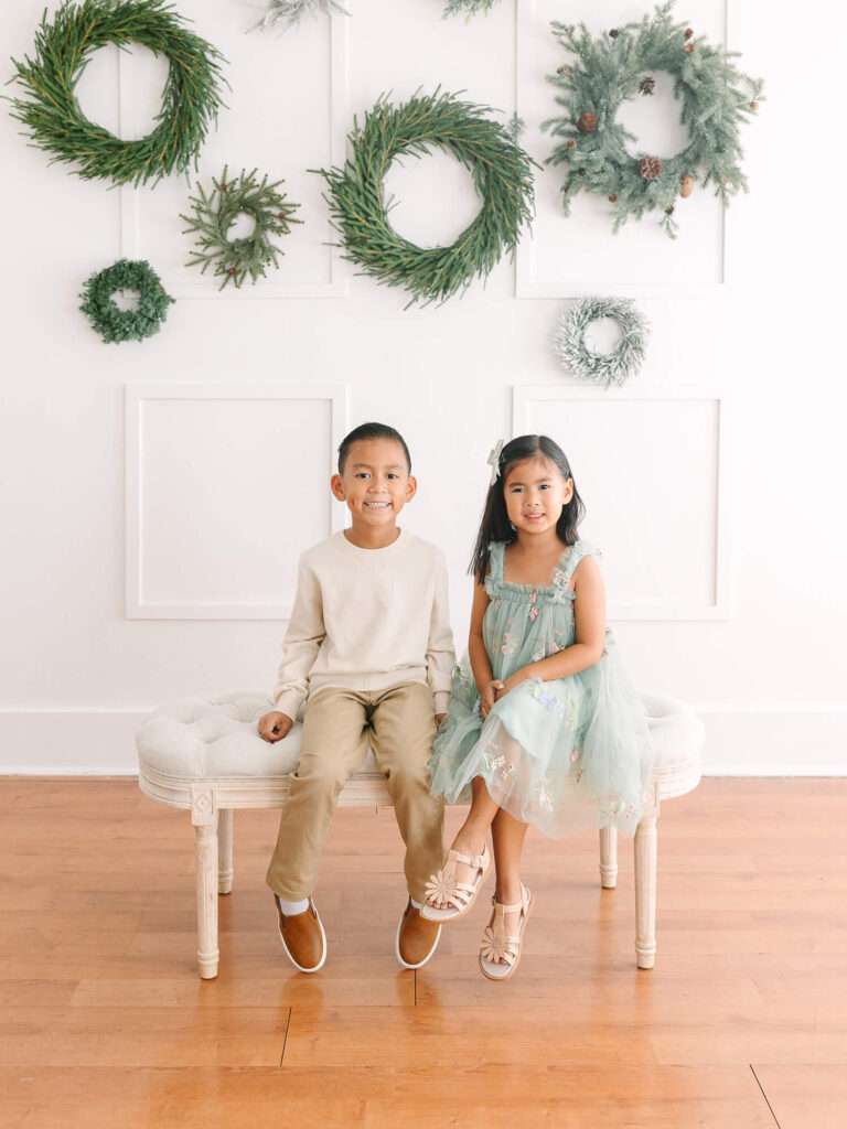 Two children sit on a beige bench; the boy wears a cream sweater, and the girl a mint dress. Green wreaths hang on the white wall behind them.