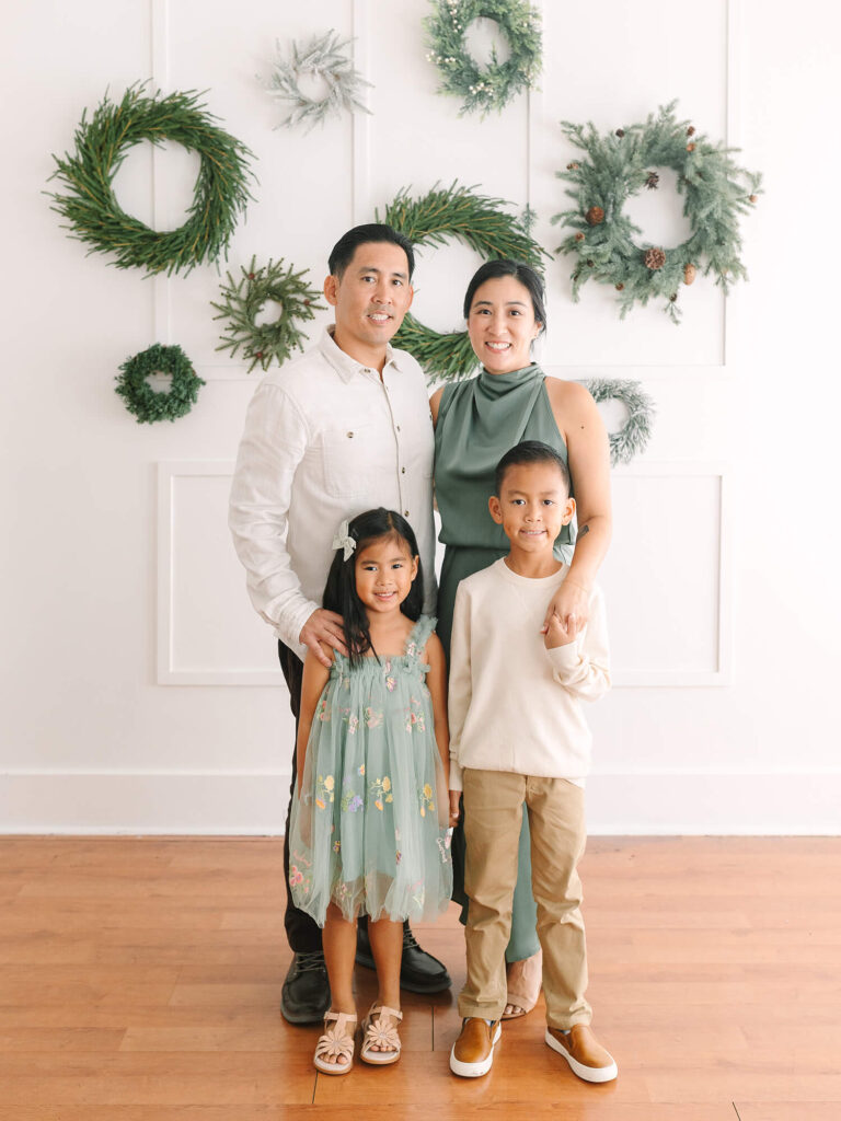 A family of four stands smiling in a festive room with green wreaths on the wall. Parents in white and green, children in pastel outfits.