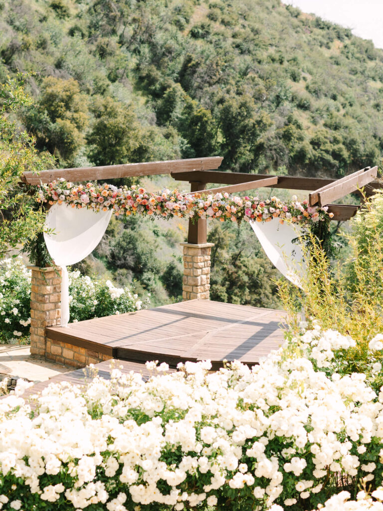 A rustic wooden pergola adorned with colorful flowers and white fabric overlooks lush, green hillside.