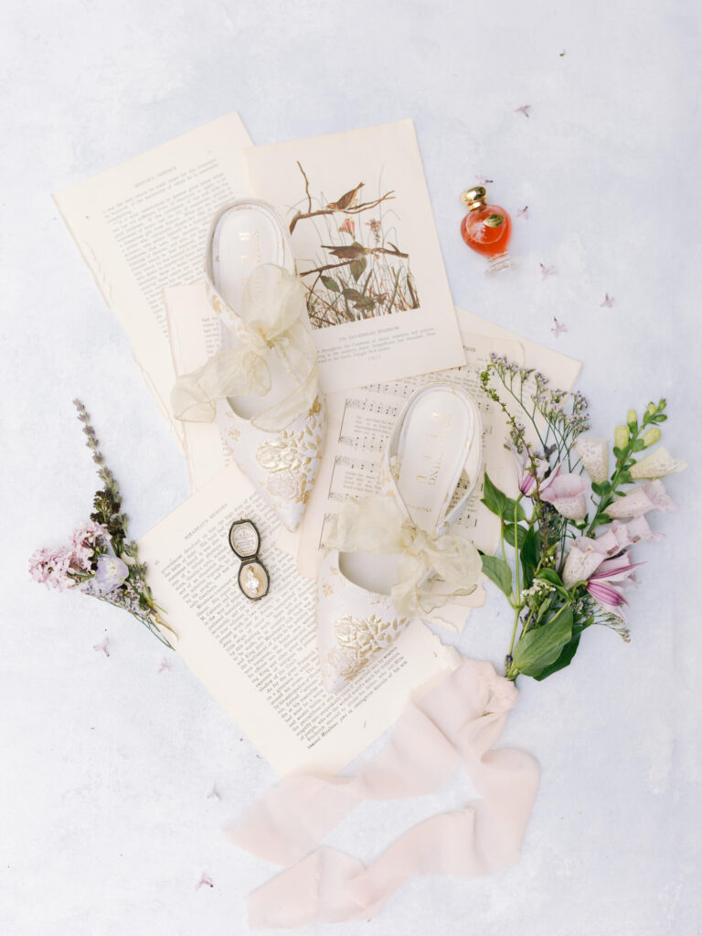 Elegant flat lay features floral shoes with bows placed on vintage book pages and sheet music, surrounded by fresh flowers, glasses, and a small perfume bottle.
