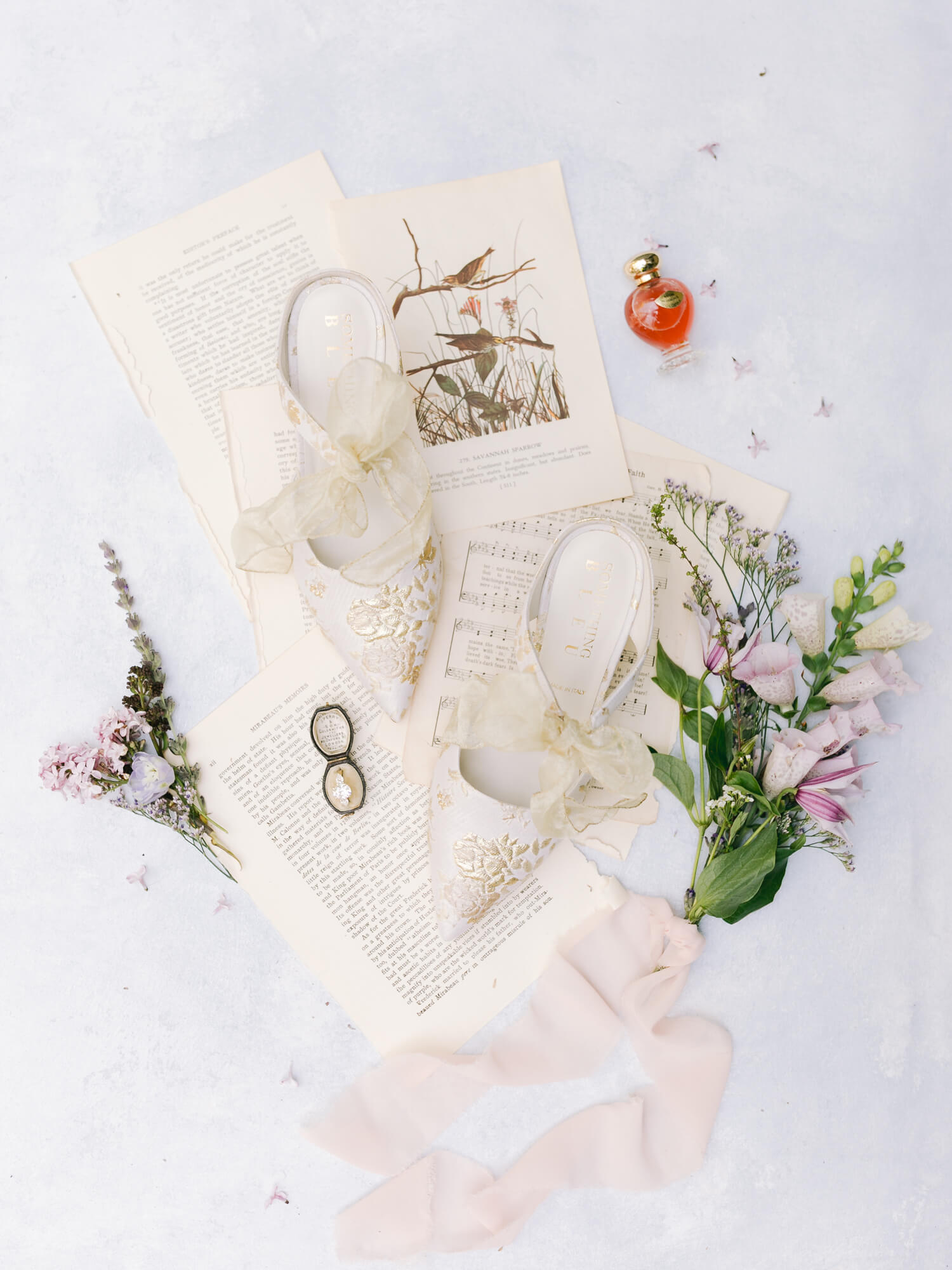 Elegant flat lay features floral shoes with bows placed on vintage book pages and sheet music, surrounded by fresh flowers, glasses, and a small perfume bottle.