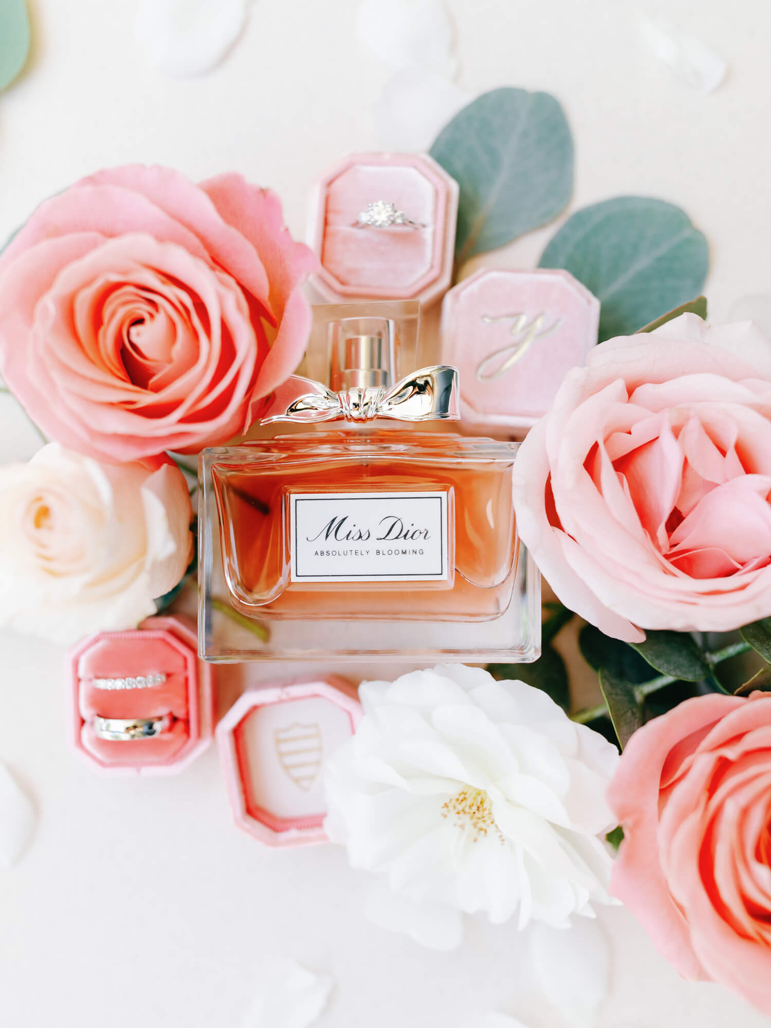 Perfume bottle labeled "Miss Dior Absolutely Blooming" surrounded by pink and white roses, placed alongside pink jewelry boxes.