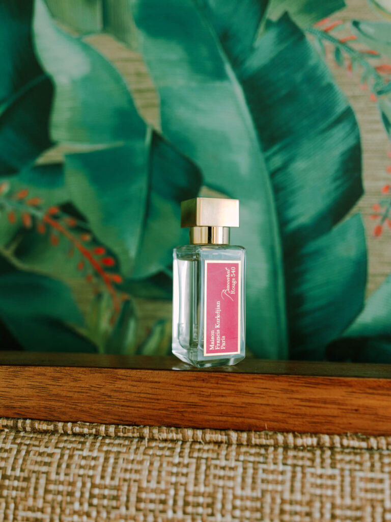 A perfume bottle with a gold cap and a pink label stands on a wooden shelf with tropical leaves in the background.